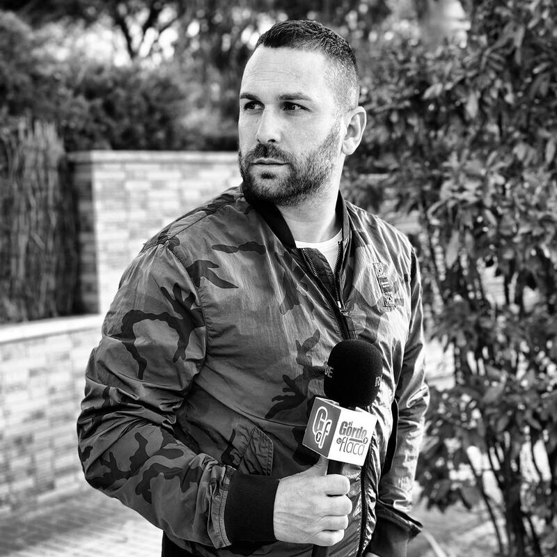 Imagen en blanco y negro de un hombre con un micrófno en la calle, mirándo al costado, con una chaqueta de camuflaje